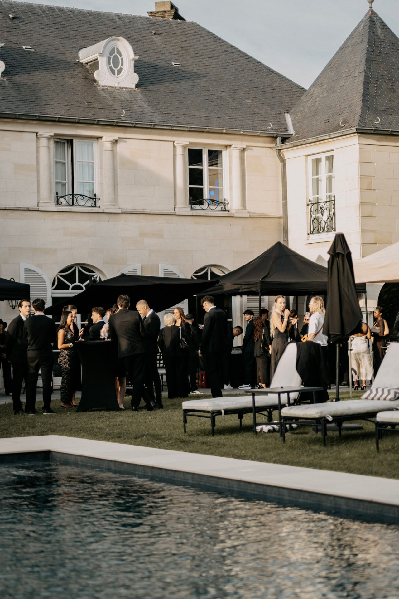 Réception en plein air sur la pelouse d'une demeure en pierre, invités en tenue de soirée près de la piscine
